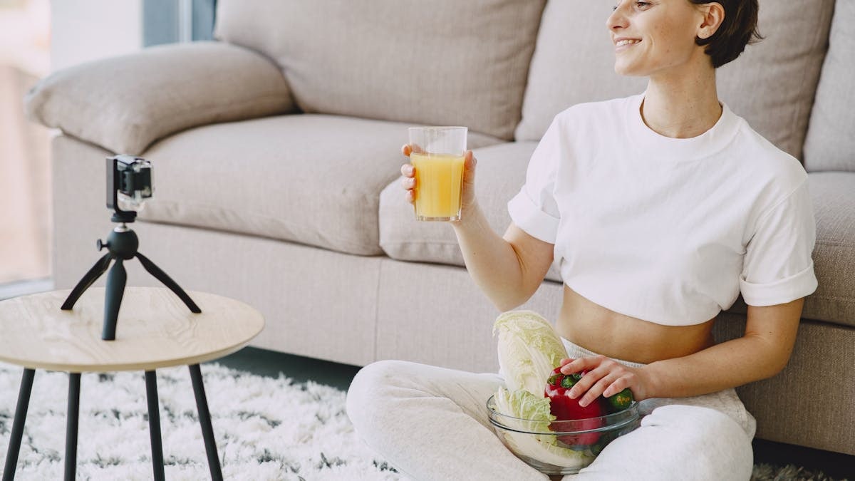 A woman smiles while holding juice and a bowl of fresh vegetables indoors.